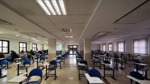 Phòng thi sáng đèn với những dãy bàn ghế đơn xếp hàng, vài thí sinh đang ngồi làm bài