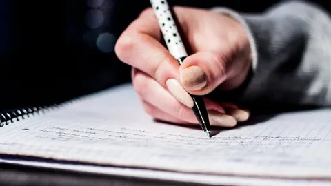 Bàn tay đang viết ghi chú bằng bút bi lên cuốn vở lò xo kẻ ô - gợi cảnh học và luyện viết tiếng Hàn