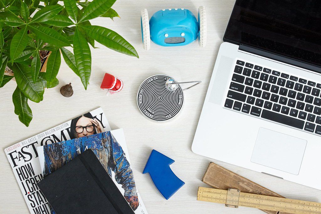 Mặt bàn nhìn từ trên xuống: laptop, cây cảnh, đồng hồ báo thức và tạp chí