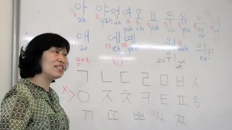 Một giáo viên đứng cạnh bảng trắng kín các nguyên âm và phụ âm bảng chữ cái Hangul trong giờ học tiếng Hàn nhập môn