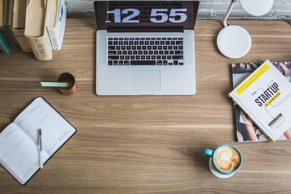 Bàn làm việc nhìn từ trên: laptop hiện đồng hồ 12:55, chồng sách, sổ tay mở có bút và tách cà phê