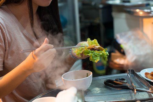 Bàn tay nâng một cuốn rau ssam còn bốc khói bên bàn nướng