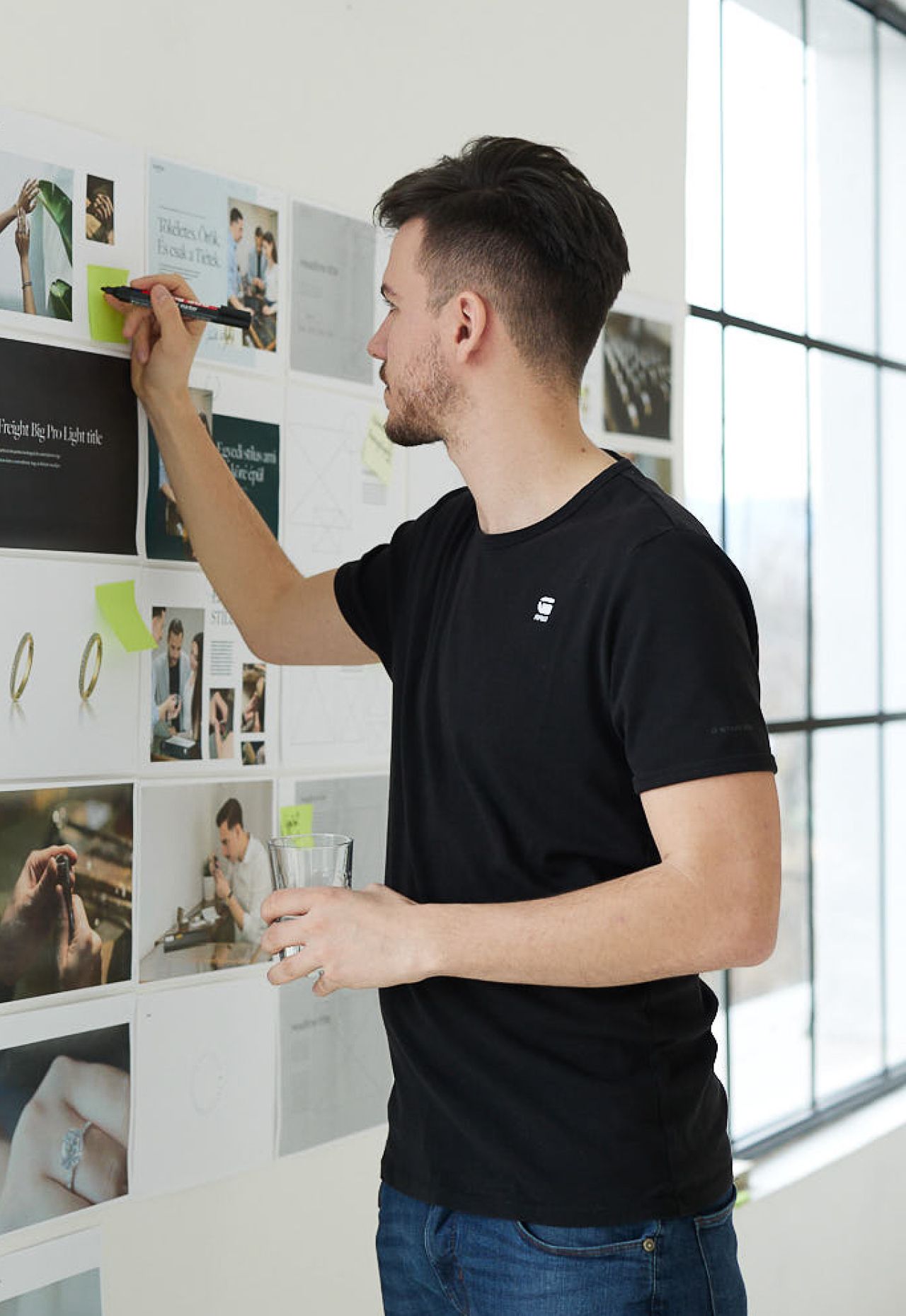 A designer annotating printed layouts pinned to the studio wall, a glass of water in hand
