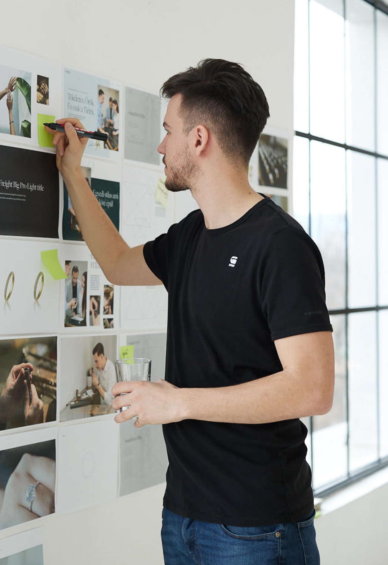A designer annotating printed layouts pinned to the studio wall, a glass of water in hand