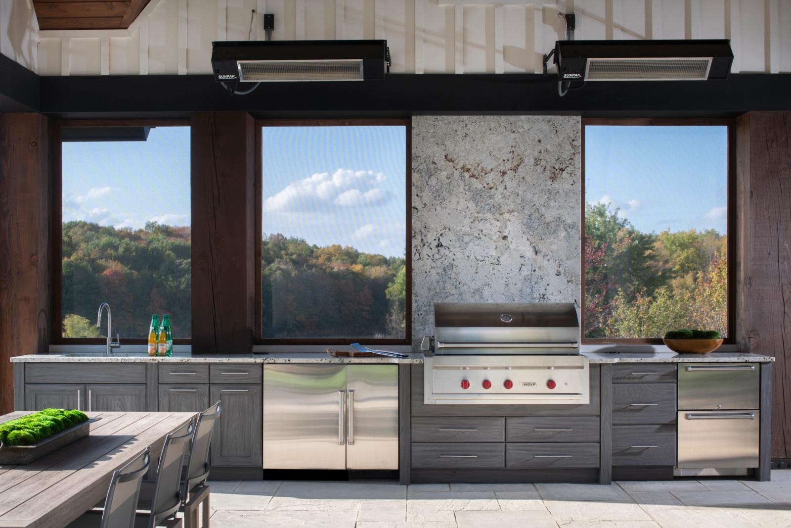 Pavilion kitchen in weathered grey cabinetry with a large built-in grill and stone slab backsplash, autumn woods beyond