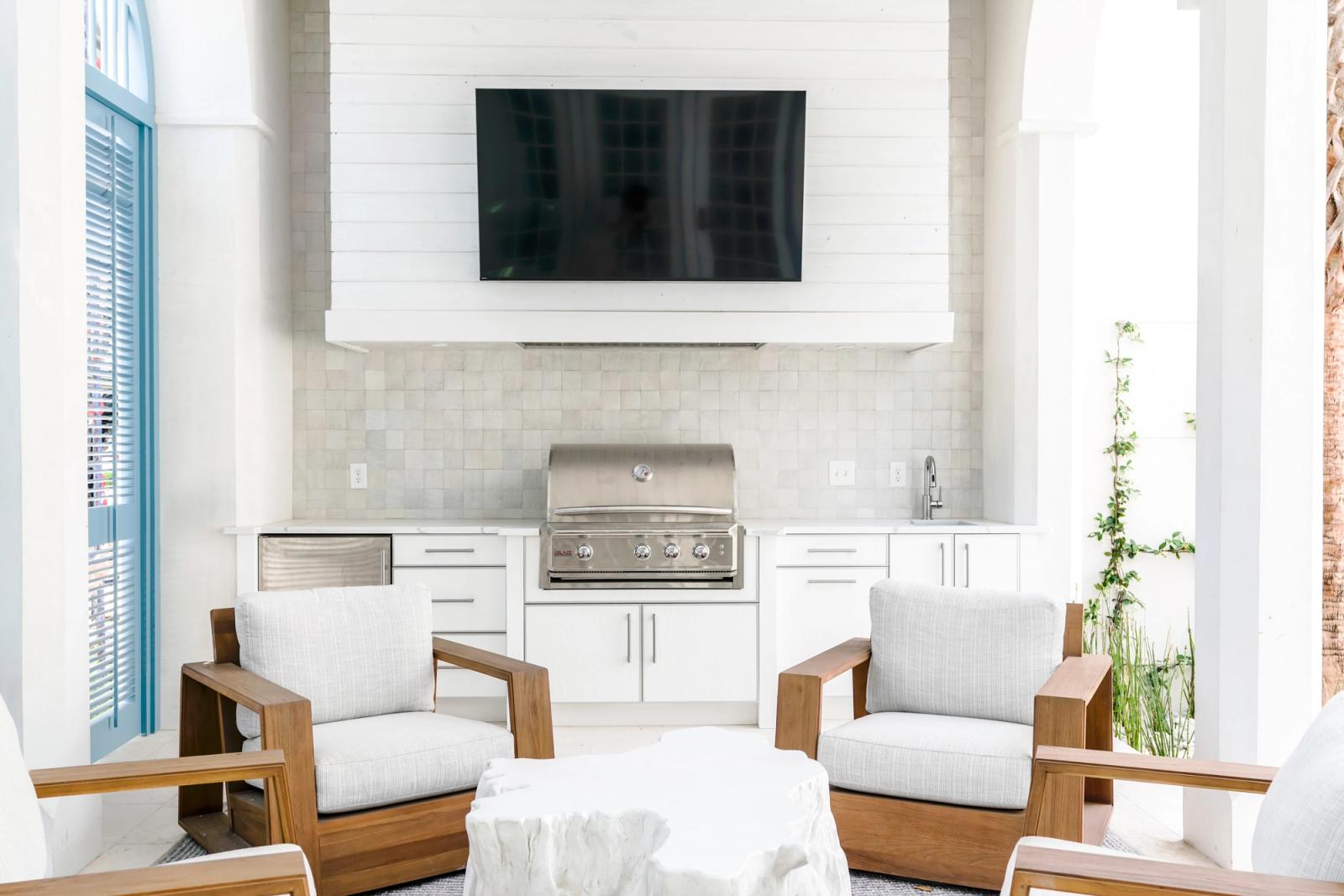 White outdoor kitchen cabinetry with a built-in grill and tiled backsplash below a wall-mounted TV, teak armchairs