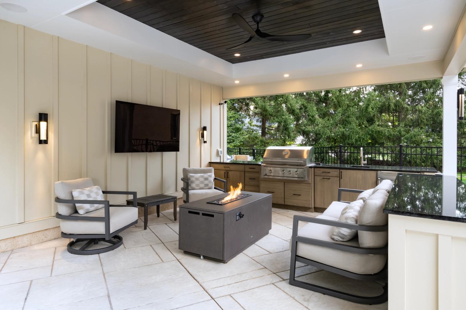 Covered porch lounge with a fire table and TV, outdoor kitchen with tan cabinetry beyond