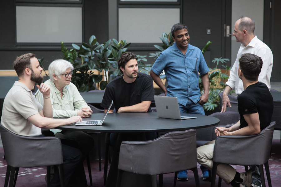 Six colleagues around a table in a plant-filled atrium, laptops open, in conversation.