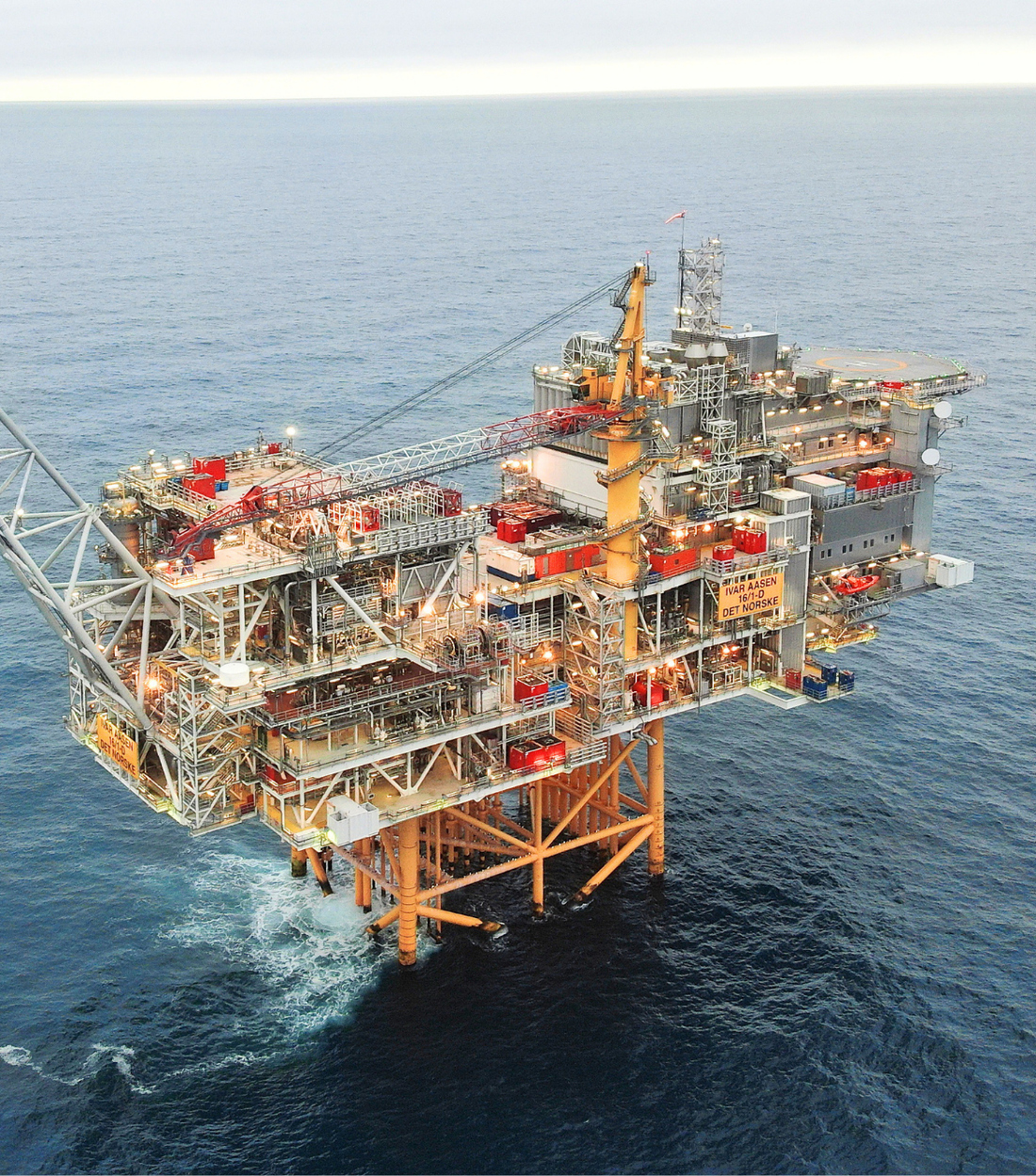 A yellow-painted offshore production platform with a long bridge out to a flare boom, gas burning at its tip, under a blue sky.
