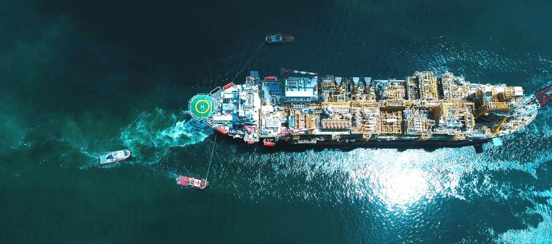 Aerial view looking straight down onto an FPSO vessel, its deck packed with process modules, tugs working alongside in turquoise water.