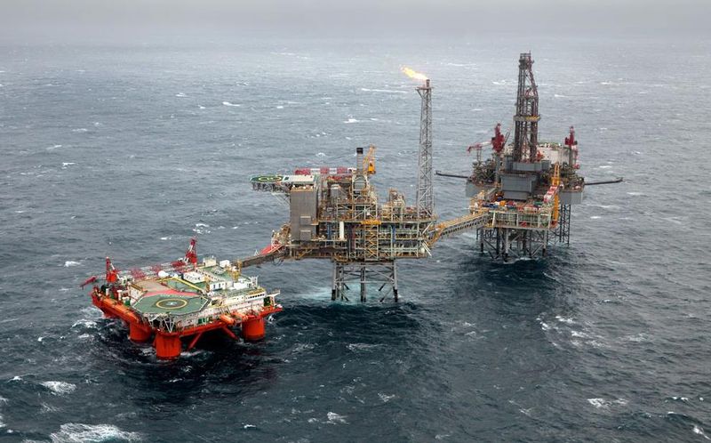 Two bridge-linked offshore platforms in grey open sea, gas burning at the top of the flare tower, with a red support vessel and helideck alongside.