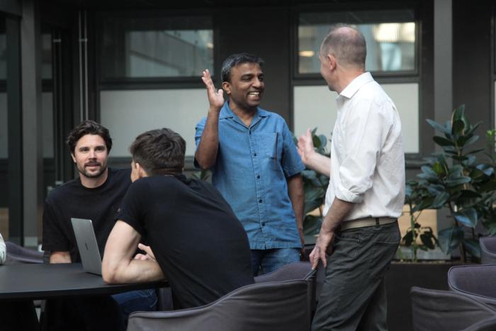 Four colleagues talking around a table in a plant-filled office atrium, one of them gesturing mid-explanation.