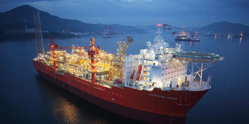 The Petrojarl Knarr FPSO lit up at dusk, moored in sheltered water below dark hills.