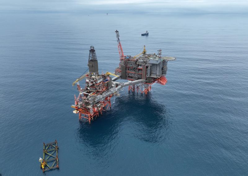 Aerial view of a bridge-linked platform complex in open sea, a drilling derrick on one platform and an unmanned jacket standing apart nearby.