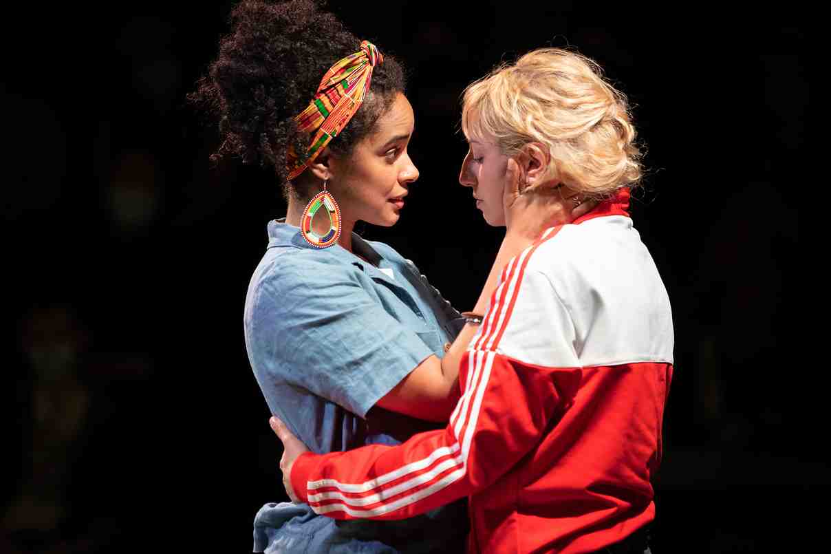 A photo of two women holding each other. On the left, a light-skinned Afro-Latina woman with dark, curly hair wears a colorful headwrap, large earrings, and a short-sleeved blue denim shirt. She cradles the face of a blonde, Near Eastern woman with blonde hair, wearing a red and white Adidas tracksuit.
