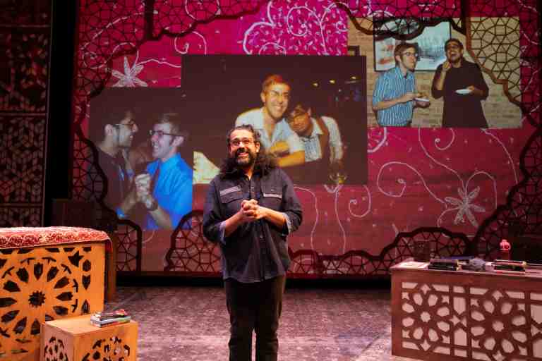 Adil Mansoor in dark blue shirt with shoulder length curly black hair standing in front of projections with a red screen in PlayCo's Ammigone