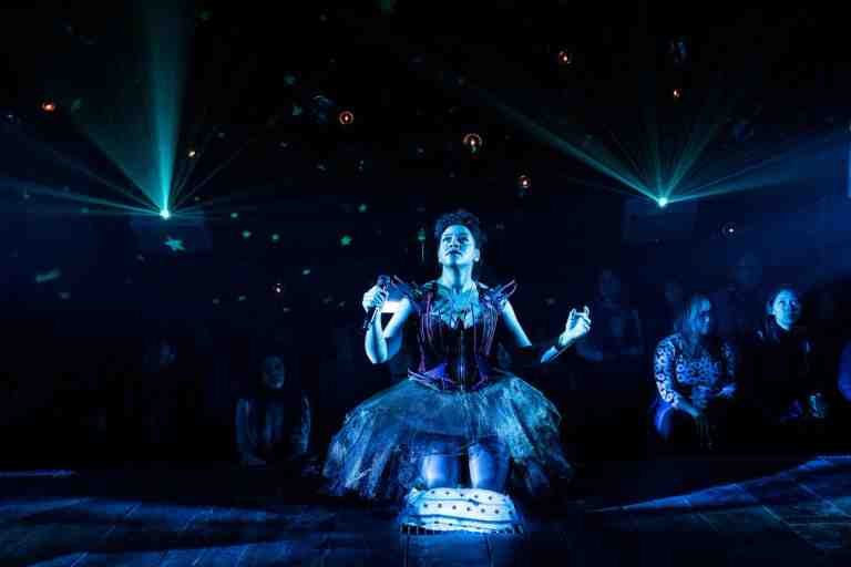 Lilli Cooper kneeling on a white patterned cushion in a dark corset dress with black and red wire embellishments and a dark translucent tulle skirt. Holding a microphone and looking up under dark greenish blue lighting. Audience visualized in background in The Wildness at PlayCo
