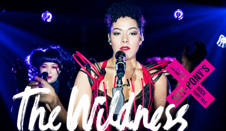 The Wildness at PlayCo title treatment. Lilli Cooper looking into camera in black corset dress with black and red wire embellishments. Back up singer behind Lilli.