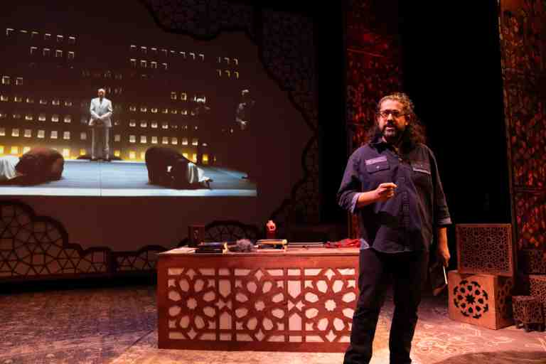 Adil Mansoor in dark blue shirt with shoulder length curly black hair standing in front of projections of an Antigone production in PlayCo's Ammigone