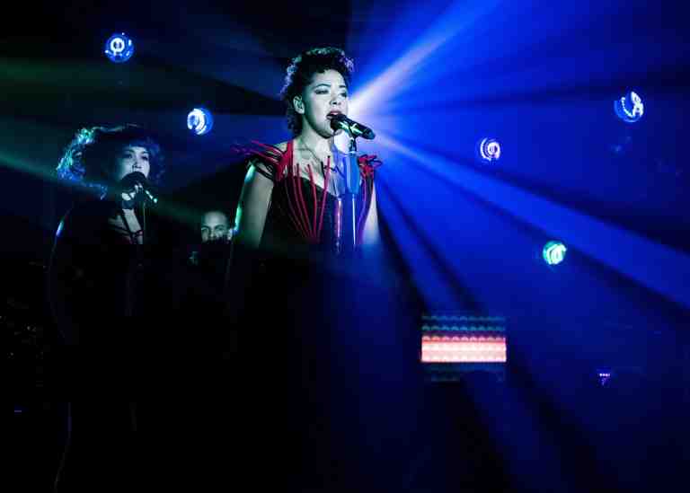 Lilli Copper singing into microphone under blue spotlight in dark corset dress with black and red wire embellishments in The Wildness at PlayCo. Backup singers and band member behind Lilli