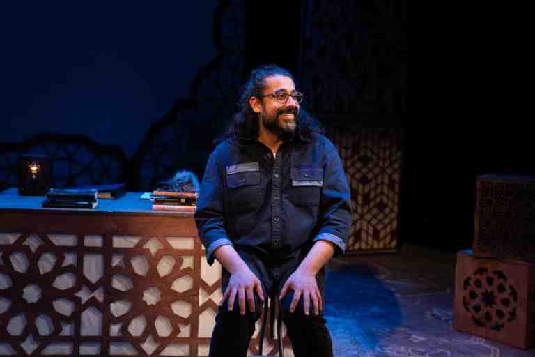 Adil Mansoor in dark blue shirt with shoulder length curly black hair smiling and sitting in front of desk in PlayCo's Ammigone