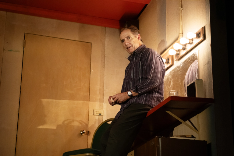 Kurt Rhoads with combed back hair in a dark button down shirt with vertical white stripes sitting on a dressing room vanity desk in Munich Medea Happy Family at PlayCo and WP Theatre