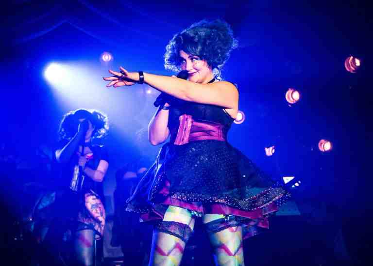 Lauren Worsham under blue hue spotlight in fantasical dress with rainbow patterned stockings and a short, curly, dark blue wig pointing to the audience. Back up singers in silhouette in The Wildness at PlayCo