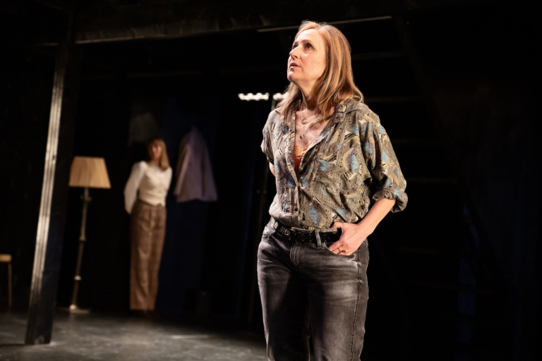 Heather Raffo standing under spotlight with blonde hair and a green patterned blouse in Munich Medea Happy Family at PlayCo at WP Theatre