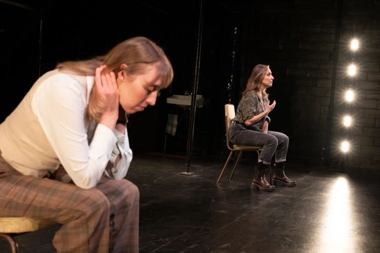 Crystal Finn in white shirt and strawberry blonde hair sitting hunched over next to Heather Raffo with blonde hair and a green patterned blouse sitting up and looking out in Munich Medea Happy Family at PlayCo and WP Theatre