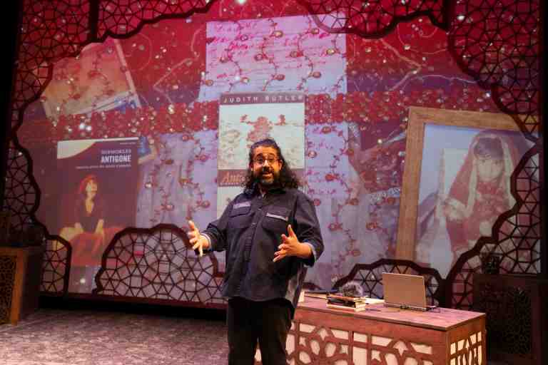Adil Mansoor in dark blue shirt with shoulder length curly blach hair and hands extended in front of projection screen of book covers in PlayCo's Ammigone