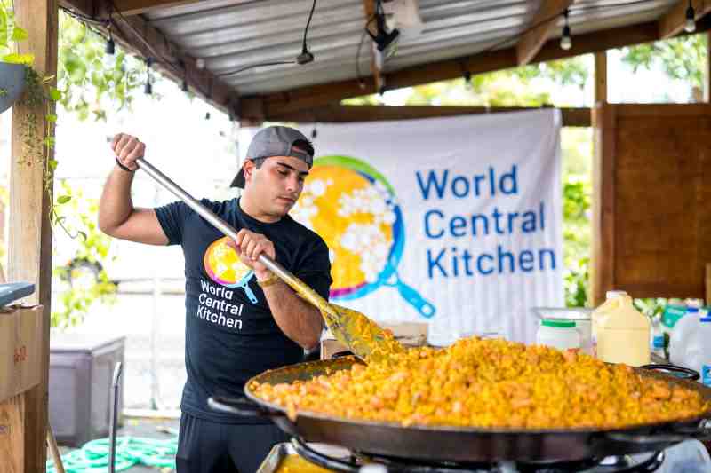A man wearing a backwards cap and a black shirt with the World Central Kitchen logo on it stirs a giant pan of paella.