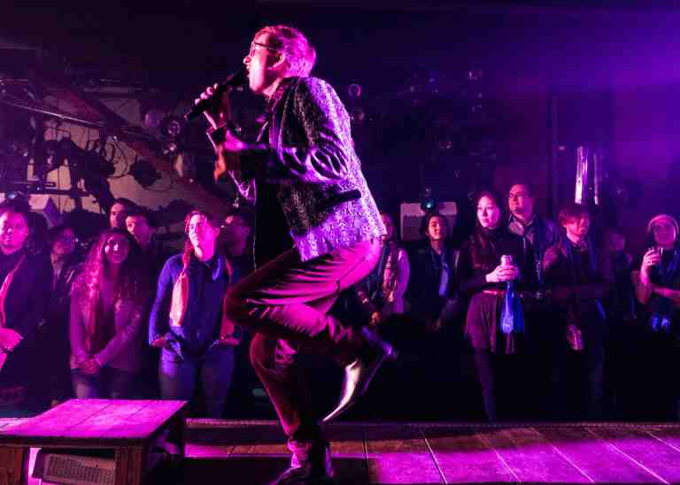 Kyle Jarrow in profile singing on stage ramp under magenta lighting with patterned tweed-like blazer, dark pants reflecting magenta light and black shoes in The Wildness at PlayCo. Audience members visualized in background.