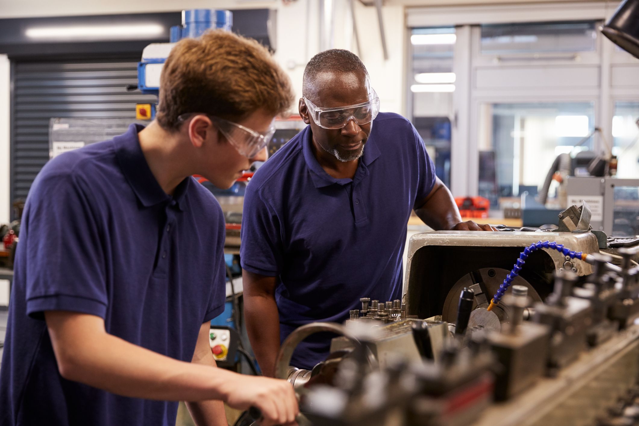 An older black man showing a younger white man how to operate a lathe