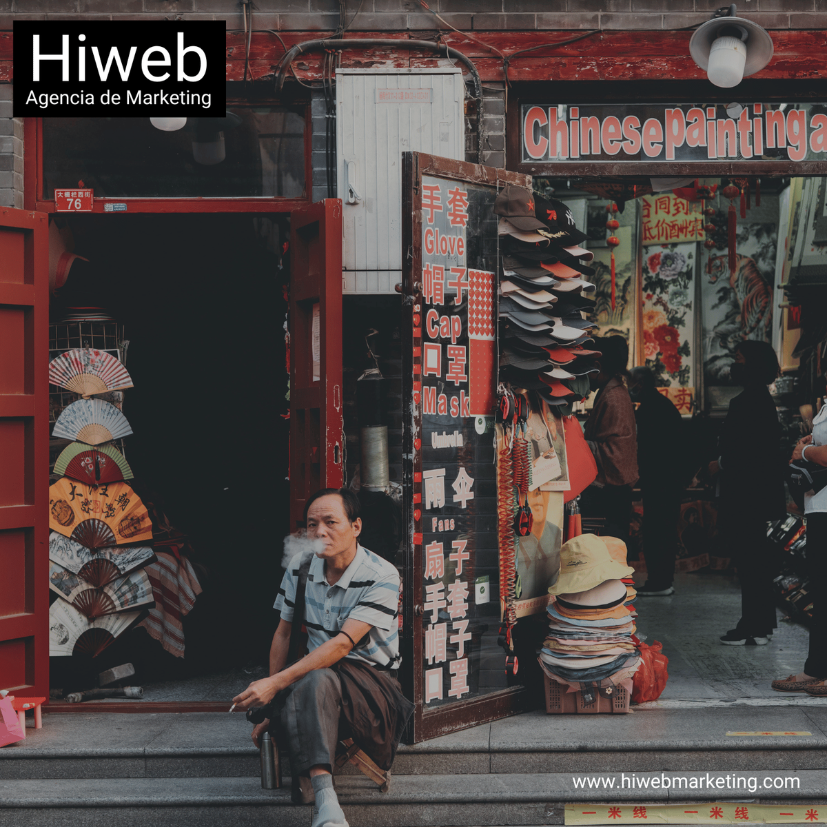 Hombre sentado fumando en la entrada de una tienda callejera tradicional con productos como sombreros y artículos a la venta, representando vida urbana y comercio local. Hiweb Agencia de Marketing.