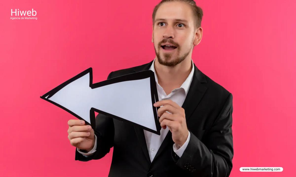 Marketing-focused image of a professional in a suit holding a large white arrow pointing left, set against a bright pink background, highlighting direction, guidance, or call-to-action messaging for business campaigns. Hiweb