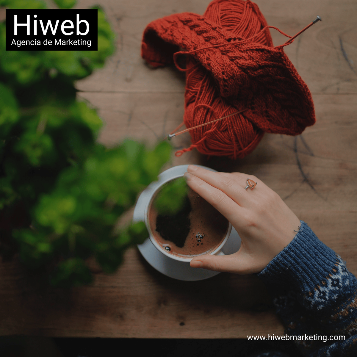 Manos tejiendo con hilo rojo sobre una mesa de madera, junto a una taza de café, representando artesanía y proceso creativo manual. Hiweb Agencia de Marketing.