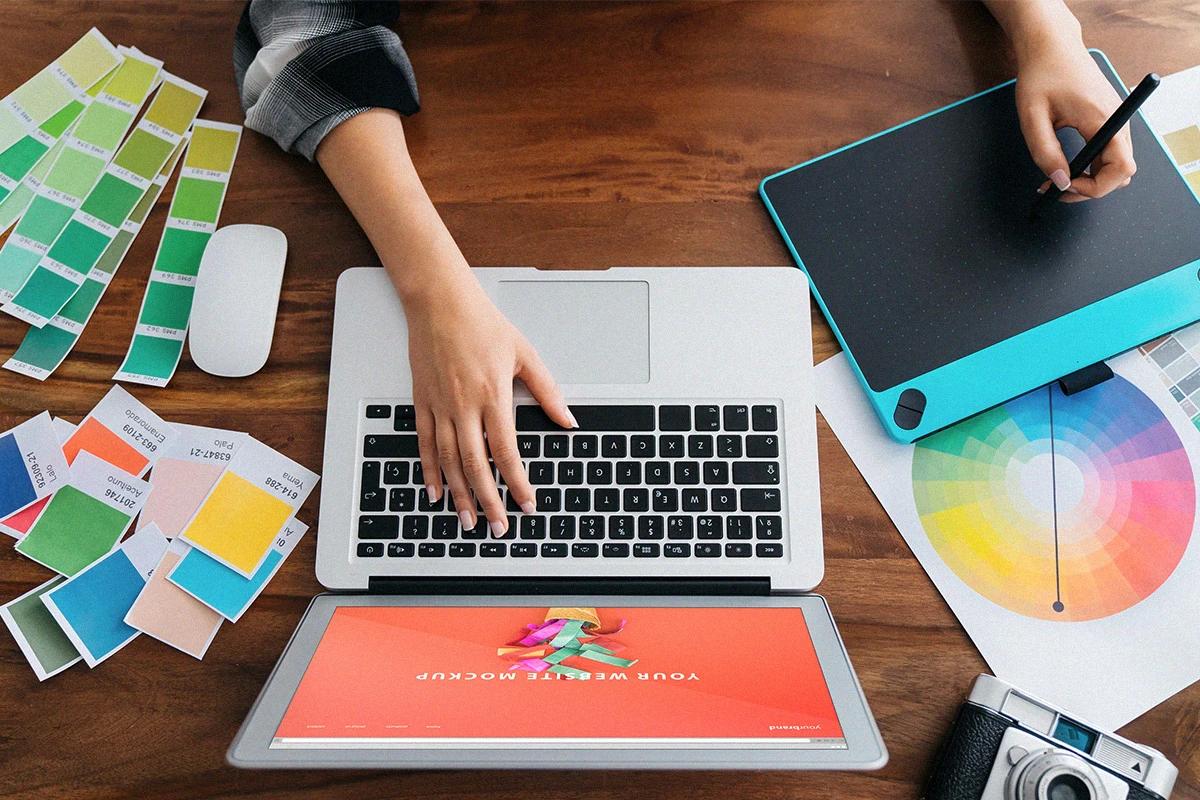 Manos trabajando sobre un escritorio con una laptop, una tableta digital y materiales de diseño gráfico como pantones o círculos cromáticos