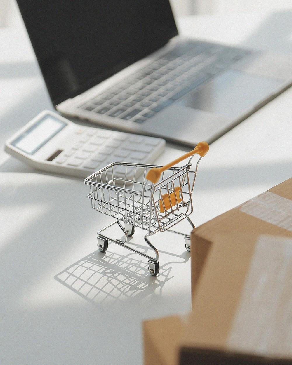 Shopping cart next to a computer and packages representing digital sales, e-commerce, and order management Hiweb.