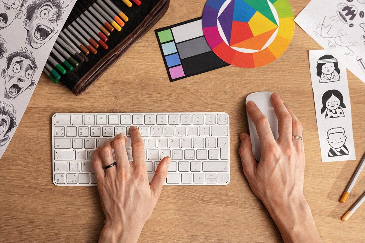 Manos sobre una mesa un teclado y mouse rodeado de elementos gráficos dibujos y colores