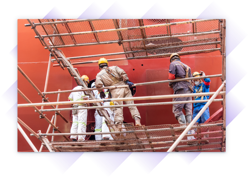 Shipyard crew in safety gear working on scaffolding beside a large vessel.