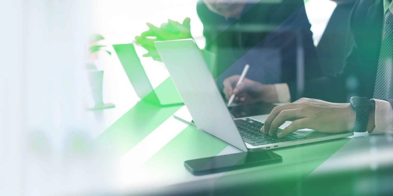 Two business professionals sitting at a desk with one typing on a laptop as explains how a TMS works and the other looks on.