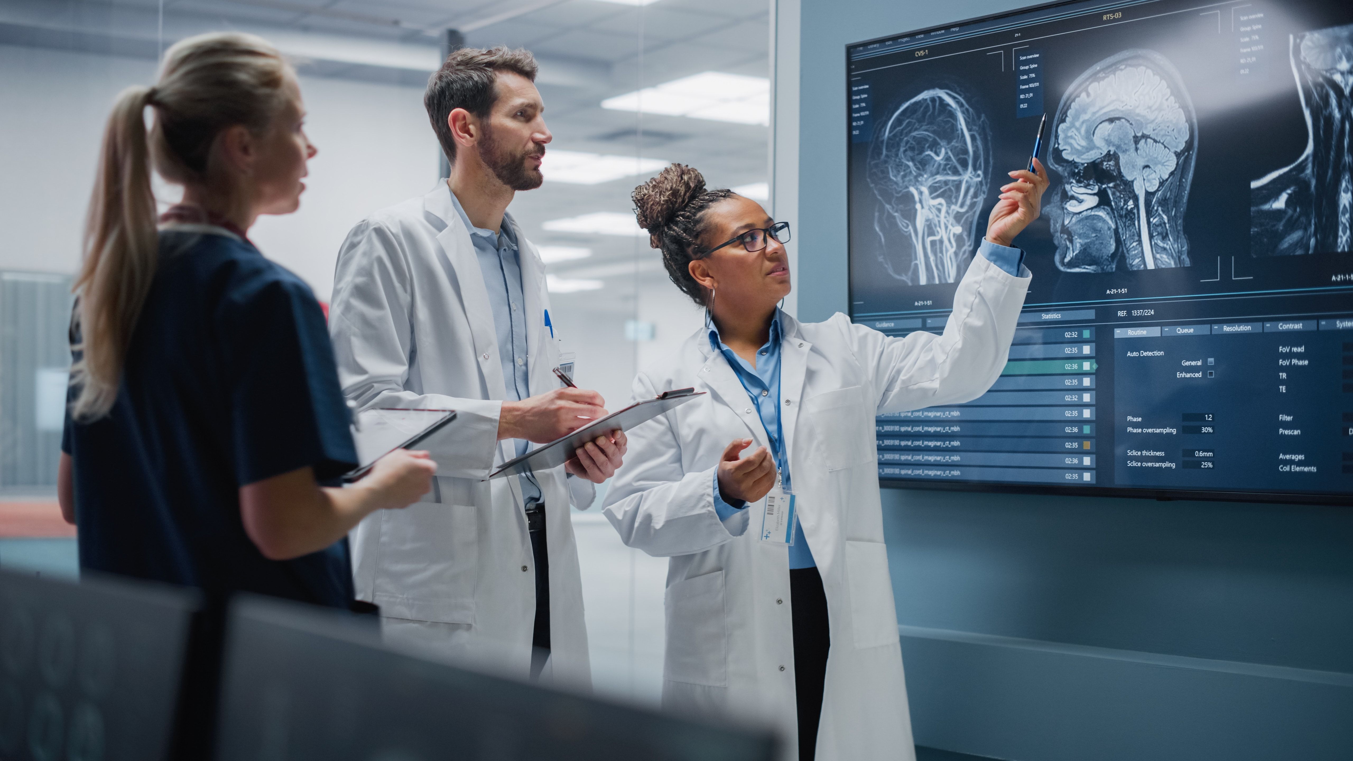 Three medical professionals in lab coats review brain scan images displayed on a large wall monitor in a clinical research setting.