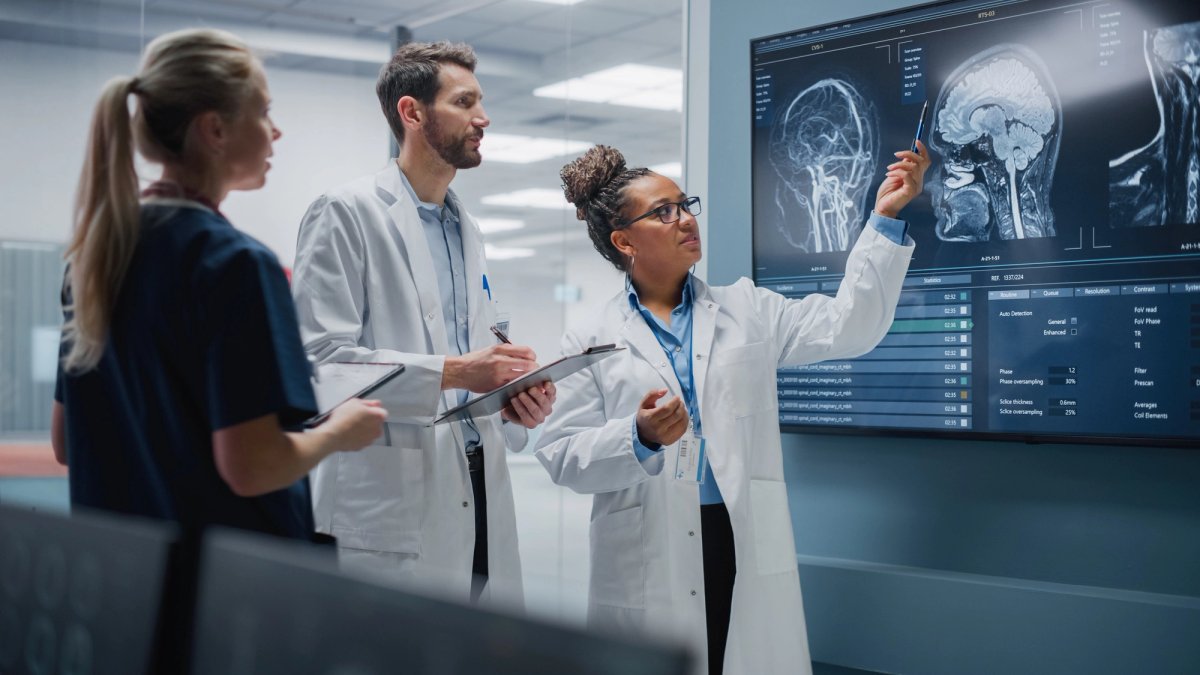 Three medical professionals in lab coats review brain scan images displayed on a large wall monitor in a clinical research setting.