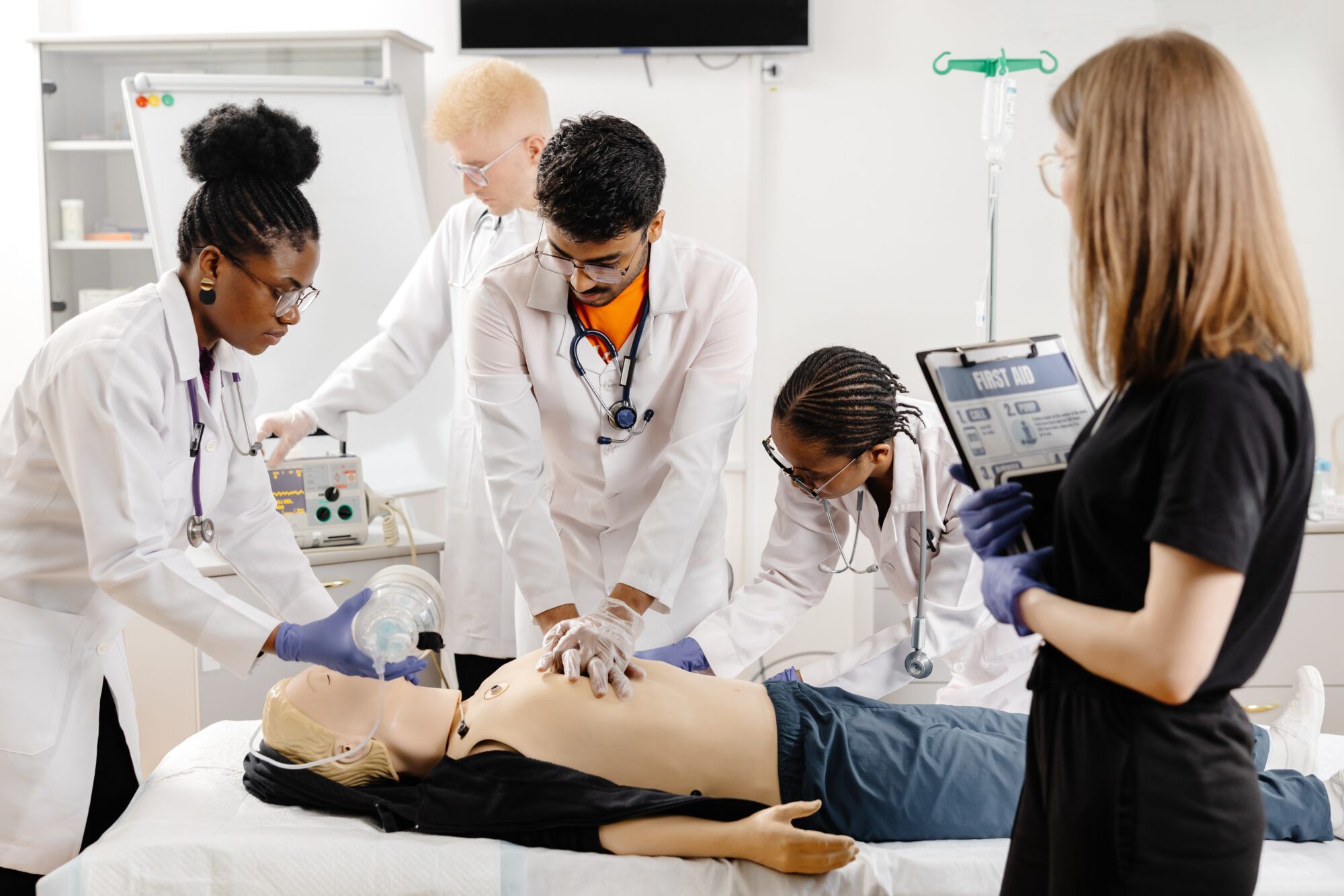 Four medical students in lab coats practice CPR and bag-mask ventilation on a training mannequin while another student holds a first-aid clipboard.