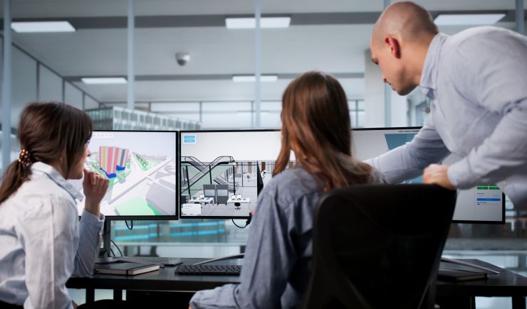 Three colleagues collaborating around dual monitors displaying technical design plans in a modern office environment.