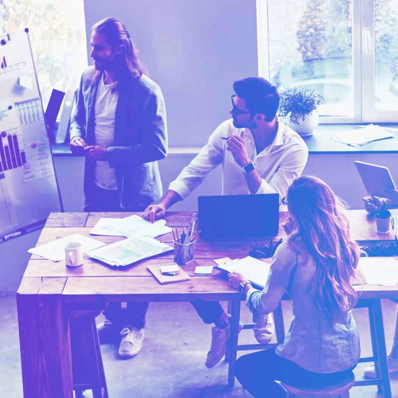 Business meeting with three professionals discussing data charts, with a blue-purple color overlay in a modern office setting.