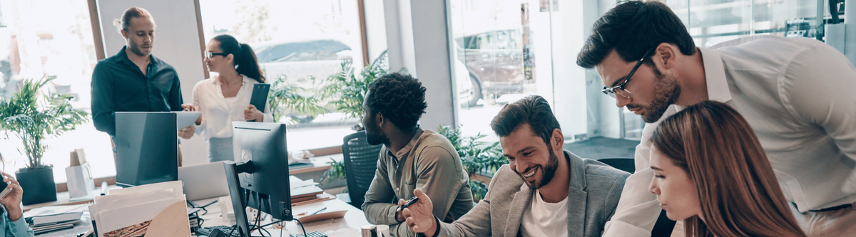 Diverse team of six professionals collaborating in a bright office with floor-to-ceiling windows overlooking a coastal cityscape.