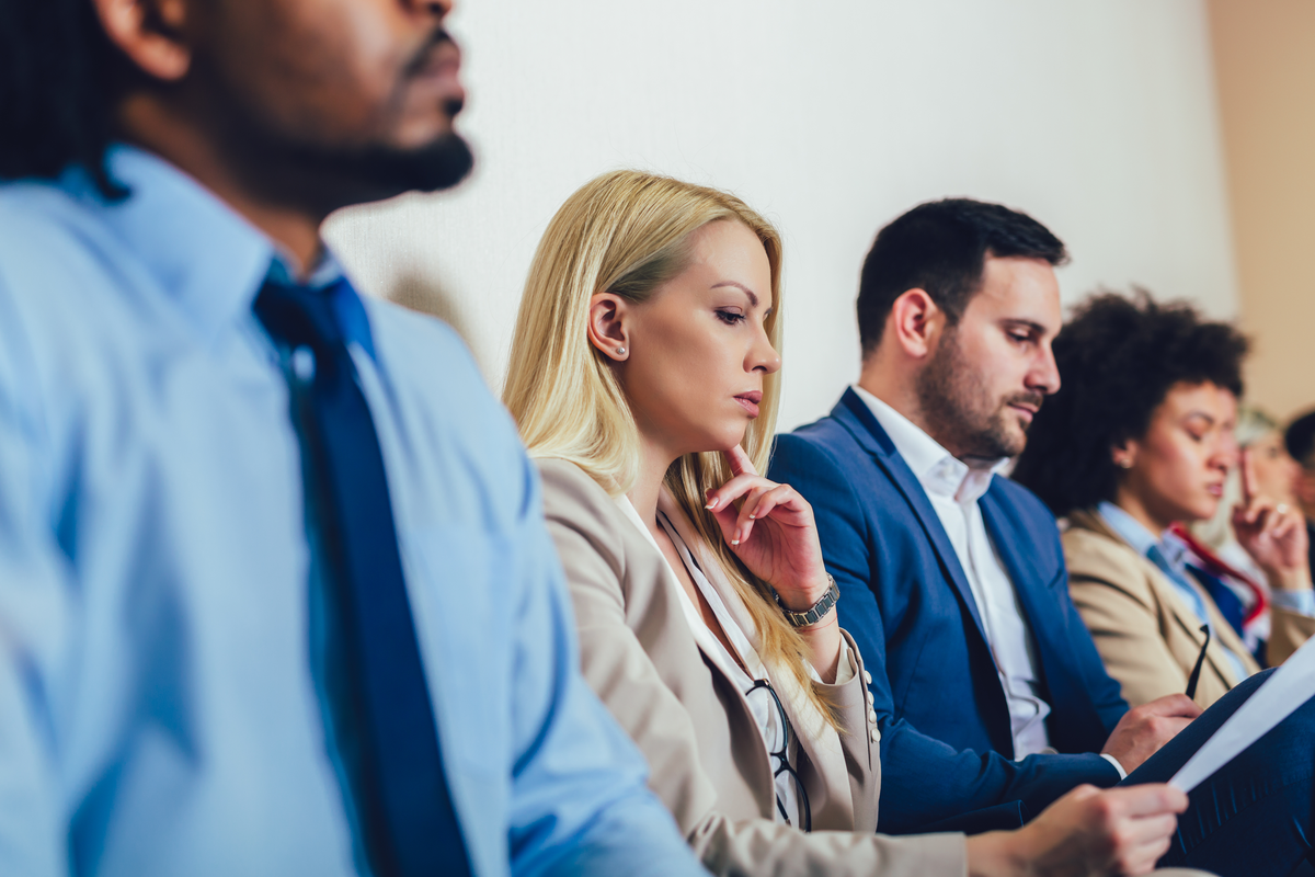 A row of people in business attire sit along a plain office wall, one resting a hand on her chin while another reviews papers.