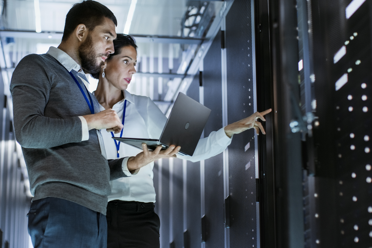 Two IT professionals stand together in a data center aisle, holding an open laptop and pointing toward a row of illuminated server racks.