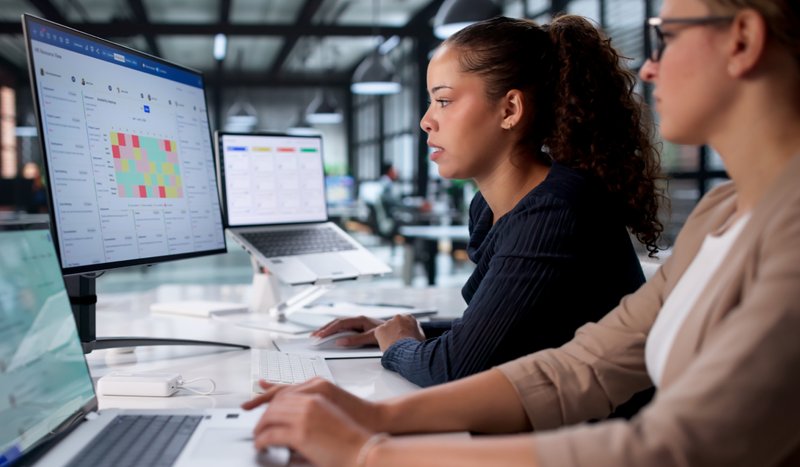 Two women at a desk in an open office plan looking at one monitor that has a treemap chart.